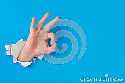 Male hand tearing through blue paper background creating ok gesture Stock Photo