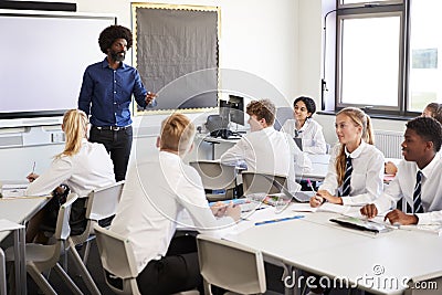 Male High School Teacher Standing Next To Interactive Whiteboard And Teaching Lesson To Pupils Wearing Uniform Stock Photo