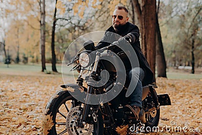 Male motorcyclist drives in nature on fast bike, wears shades, black jacket, gloves, jeans and boots, enjoys autumn season, spends Stock Photo