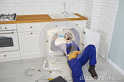 Male plumber in workwear repairs and maintains siphon under sink in kitchen of apartment. Stock Photo