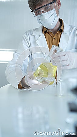 Male scientist wearing white protective gloves, mask and glasses is holding a yellow chemical solution inside an Stock Photo