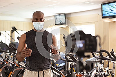 Male sports instructor in gym with a face mask recording video content for online classes, streaming Stock Photo