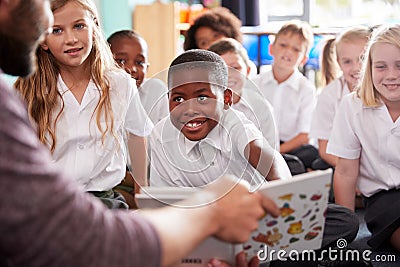 Male Teacher Reading Story To Group Of Elementary Pupils Wearing Uniform In School Classroom Stock Photo