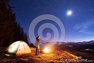 Male tourist have a rest in his camp at night, near campfire and tent under night sky full of stars and the moon Stock Photo