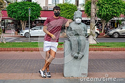 A male tourist wearing a medical face mask poses near a stone sculpture of a masked accordionist on Ataturk Boulevard in Alanya, Editorial Stock Photo