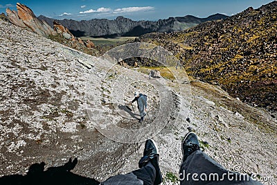 Male Traveler Sits On Top Mountain And Enjoys Mountain View In Summer. Group Of Tourists Climbs Uphill. Point Of View Shot Stock Photo