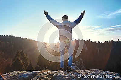 Male traveler standing on the cliff against wooded hills and cloudy sky at sunrise. Man stands hands up, greeting the Stock Photo