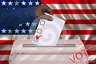 Male voter drops a ballot in a transparent ballot box against the background of the national flag of America, concept of state Stock Photo