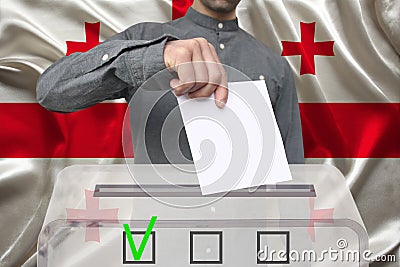 Male voter drops a ballot in a transparent ballot box against the background of the national flag of Georgia, concept of state Stock Photo