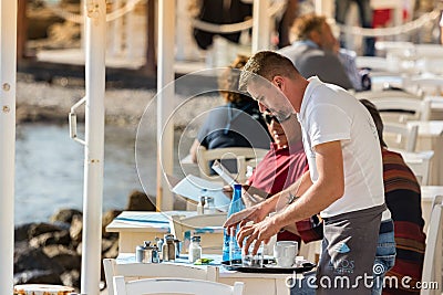 A male waiter clearing a table outdoors at a greek tavern in the waterfront of Mykonos, Greece Editorial Stock Photo