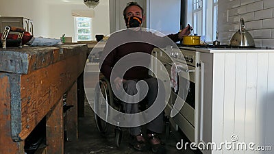 Male in a wheelchair cooking food in the kitchen. He is wearing a face mask Stock Photo