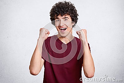 Male winner with curly hair, clenches fists and exclaims with happiness, dressed in casual outfit, celebrates his victory or trium Stock Photo