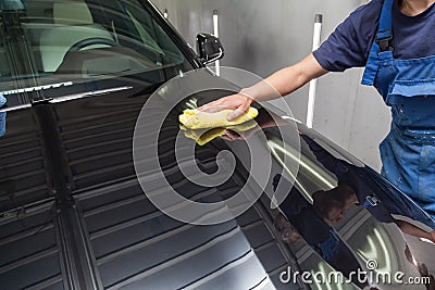 A male worker in blue overalls wipes the hood of a black car with a yellow microfiber cloth during polishing before applying a Stock Photo