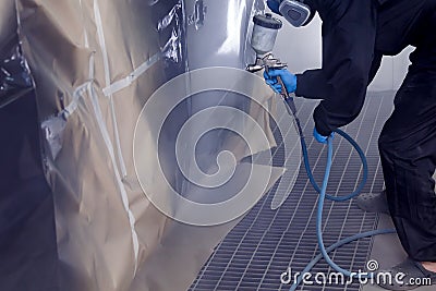 A male worker in jumpsuit and gloves paints with a spray gun a front frame part of the car body in black after being damaged at an Stock Photo