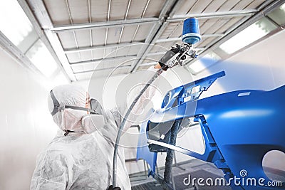 Male worker in protective clothes and mask painting car bumper using spray paint. Stock Photo