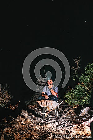 A male worker is sitting in a wheelbarrow. Night view. Headlamp illuminates the trail. The concept of heavy utility and Stock Photo