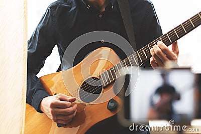 Man with acoustic guitar in a shirt and cap plays live staying home for the audience in front of the smartphone camera during Stock Photo