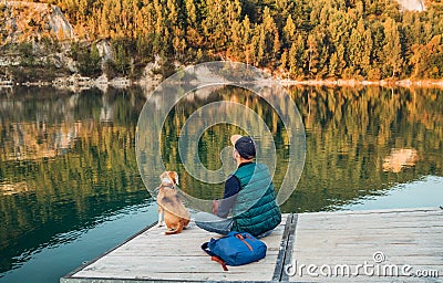 Man as a dog owner and his friend beagle dog are sitting on the wooden pier on the mountain lake and enjoying the landscape during Stock Photo