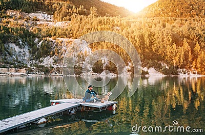 Man as a dog owner and his friend beagle dog are sitting on the wooden pier on the mountain lake and enjoying the landscape during Stock Photo