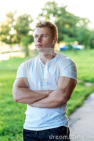 The man atlen stands in the park with headphones, a white T-shirt. Summer lifestyle, motivation is strong. Muscular arms Stock Photo