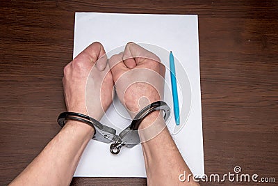 A man with bare hands in handcuffs sits at a table in front of a blank sheet of paper and a fountain pen. 4 Stock Photo