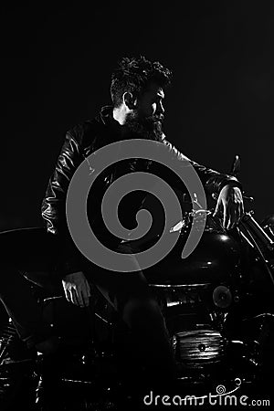 Man with beard, biker in leather jacket lean on motor bike in darkness, black background. Brutality and masculine Stock Photo