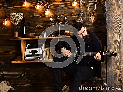 Man with beard holds black electric guitar. Guy in cozy warm atmosphere play music. Man bearded musician enjoy evening Stock Photo