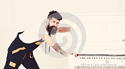 Man with beard and mustache, worker in overalls pushes piano, white background. Courier delivers furniture in case of Stock Photo
