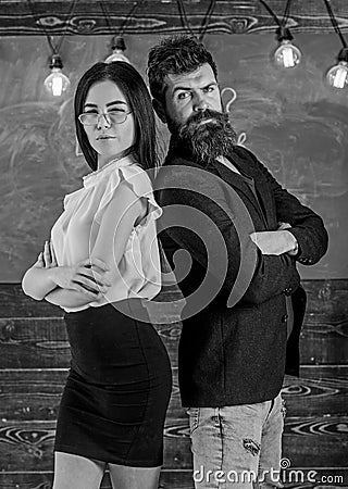 Man with beard and teacher in eyeglasses stand back to back, chalkboard on background. School staff concept. Lady and Stock Photo