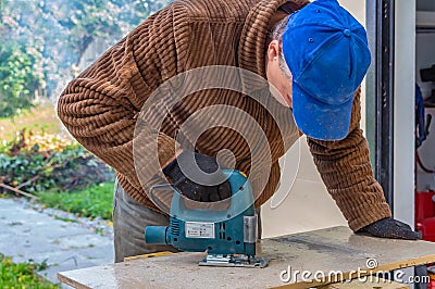 A man in the black working gloves and a brown jacket and a blue hat cuts a board using a Jigsaw power tool on a work Stock Photo