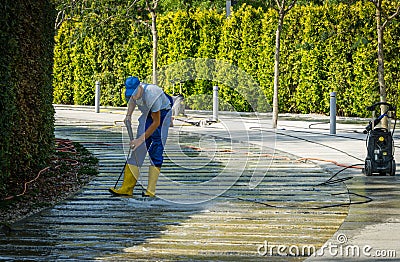 Man in blue overalls and yellow boots cleans granite bed of artificial fast river with car wash. Public landscape city park Krasno Editorial Stock Photo