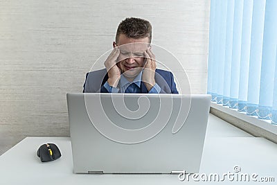 man in a blue suit with a sore head in the office. The concept of overwork at work. A tired businessman holds his head Stock Photo
