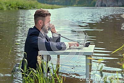 A man in a business suit is sitting at a white table in a swamp, is looking at a laptop and talking on the phone Stock Photo