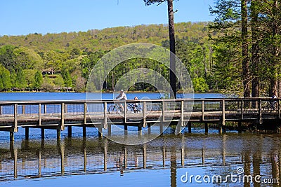 A man and a child riding bikes across a long brown wooden bridge over a rippling blue lake surrounded by lush green trees Editorial Stock Photo