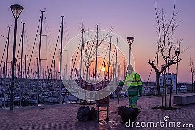 Man is cleaning up the city in the morning near a marina filled with several sailboats Editorial Stock Photo