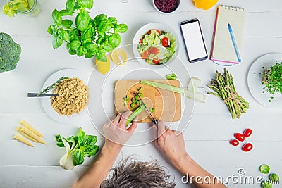 Man cutting celery and cooking vegan fresh salad with vegetables. Blank screen smart phone and notebook for daily diet planning. Stock Photo