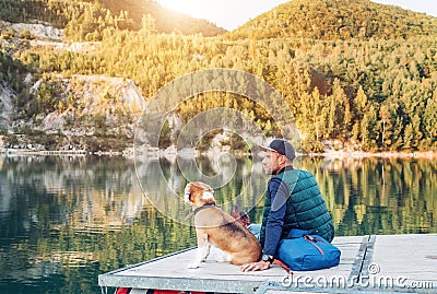 Man dog owner and his friend beagle dog are sitting on the wooden pier on the mountain lake and enjoying the landscape during Stock Photo