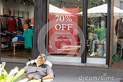 Man falls asleep in outdoor chair in front shop window and discount sign waiting for shopping wife Editorial Stock Photo