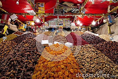 Man in a food stall holding out scoop full of nuts at camera surrounded by vibrant colored fruits and nuts Editorial Stock Photo
