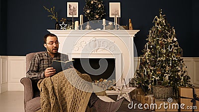 A man in glasses in chair reading a book at home at Christmas time out loud. Christmas iterior. Stock Photo