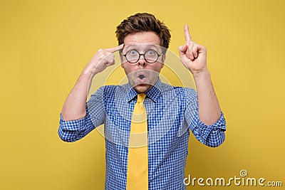 Man in glasses yellow tie and blue shirt has intriguing expression pointing finger up having great idea looks happily Stock Photo