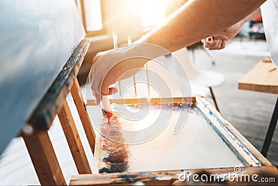 Man hand of artist closeup squeezing paint from a tube on a palette. Painter mixes oil paint colors on the palette in workshop Stock Photo