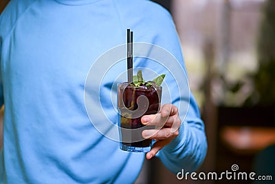 Man hand holding a drink in a tall glass. Refreshing cocktail with mint and plastic straw. Person in blue sweater. Stock Photo