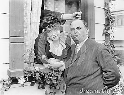 Man having his hair pulled by a hand coming out of a window while standing next to a woman Stock Photo