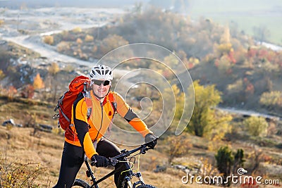 Man in helmet and glasses, with backpack stay on the bicycle under landscape with rocks and hill. Stock Photo