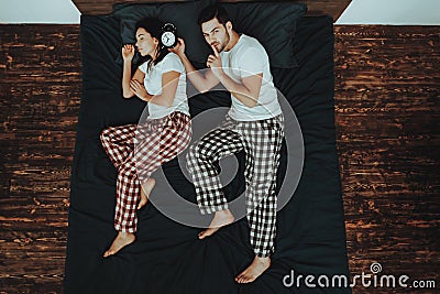 Man is Holding Alarm Clock near Woman`s Ear on Bed Stock Photo