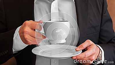 Man holding a cup of coffee with cream and small white plate, dressed in white dress shirt and black suit and wearing a watch Stock Photo