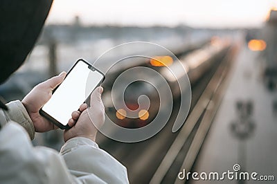 The Man is holding a mock up smartphone in his hand, against the background of the train at the railway station. Stock Photo