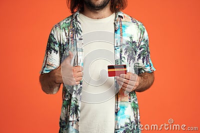 A man holds a credit card and shows thumbs up close up. Male traveler with a credit card in the studio on an orange Stock Photo