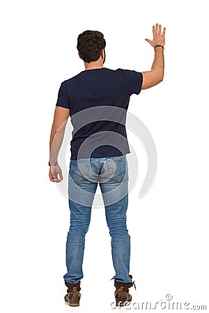 Man In Jeans And Blue T-shirt Is Standing With Arm Raised And Waving Hand. Rear View Stock Photo